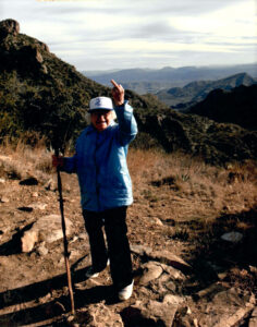 Woman in blue coat and white baseball cap holds a walking stick in one hand and holds the other hand up with the third finger extended. The Mountains of Big Bend are in the background.