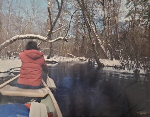 Woman in warm jacket paddles in front of a canoe on a stream. The banks of the stream and the trees are covered with snow.