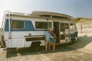 Joe and Holly Lucas stand next to their Winnebago recreational vehicle that has a canoe on top and is parked near sand dunes.