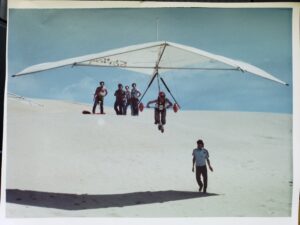 Person holding onto support bars under a big hang-gliding sail above the sand dunes.