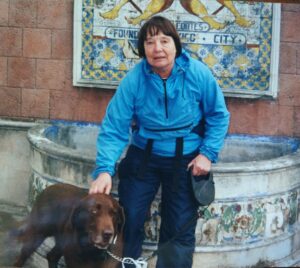 Holly Lucas in blue windbreaker, sits on the rim of a fountain, petting her brown dog, Griz
