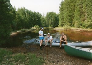 A woman is standing, while two men sit on the gunnel of a canoe that is on the shore in the Jersey Pine Barrens.
