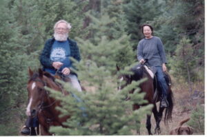Joe and Holly ride horses on a trail through the woods in Montana